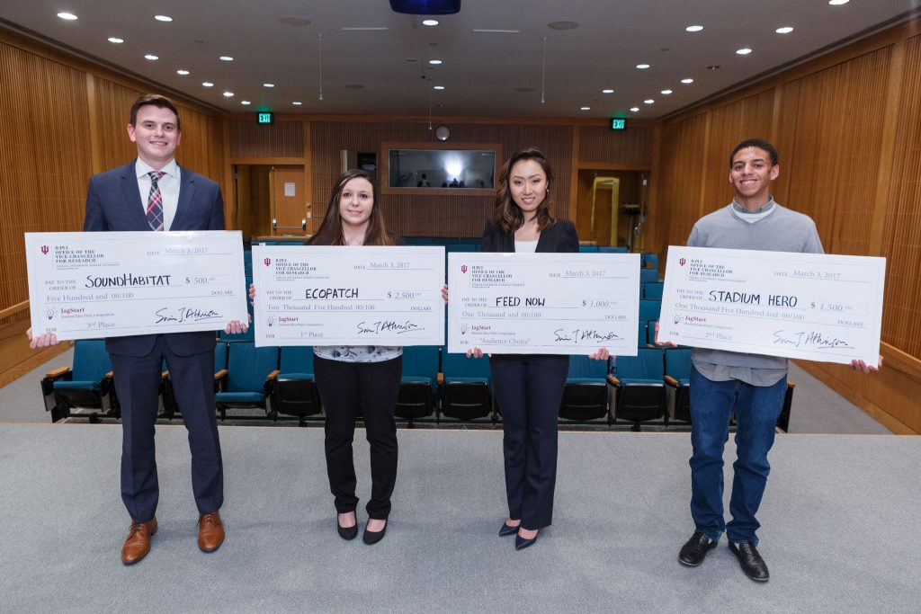 Four students, two women and two men, stand shoulder to shoulder, holding oversized checks. The students were named winners of the 2017 JagStart Business Pitch competition held at IUPUI.