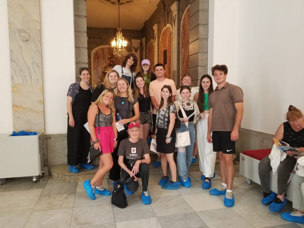A group of students smiling at the camera in a hallway of the Royal Palace. They wear blue shoe coverings to protect the floor