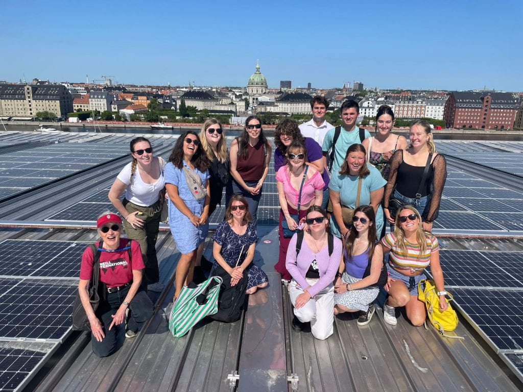 A group of students and professors smile in the sunshine on the roof of the Danish opera house. Solar panels surround them with a flat, densely packed row of buildings in the background