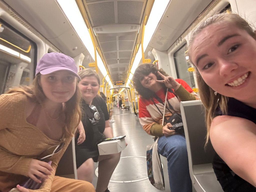 Four students smile in a selfie taken inside a grey and yellow subwait train