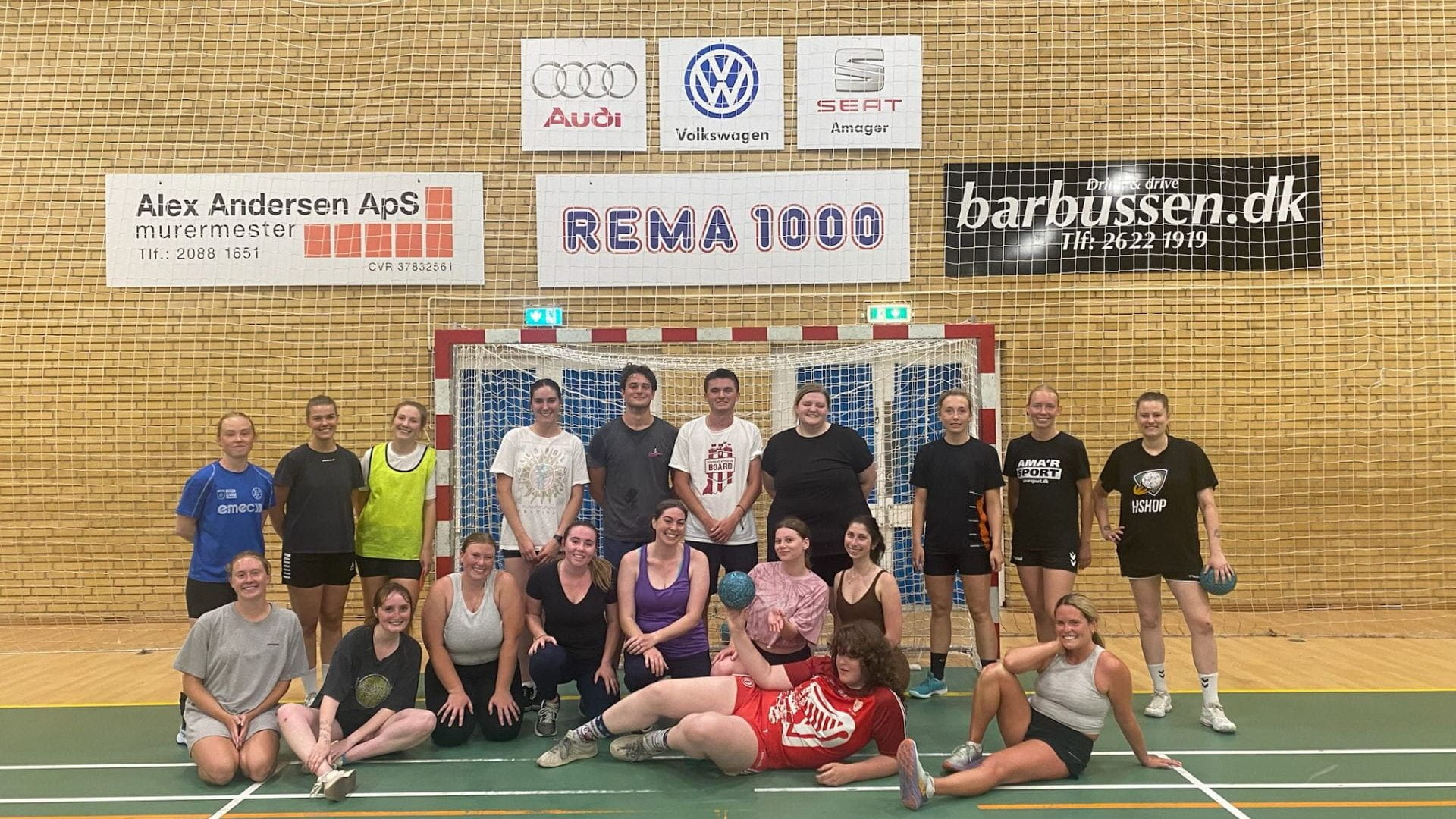 A group of students and handball players pose on a green handball court in front of a net and a brick wall with sports banners behind them