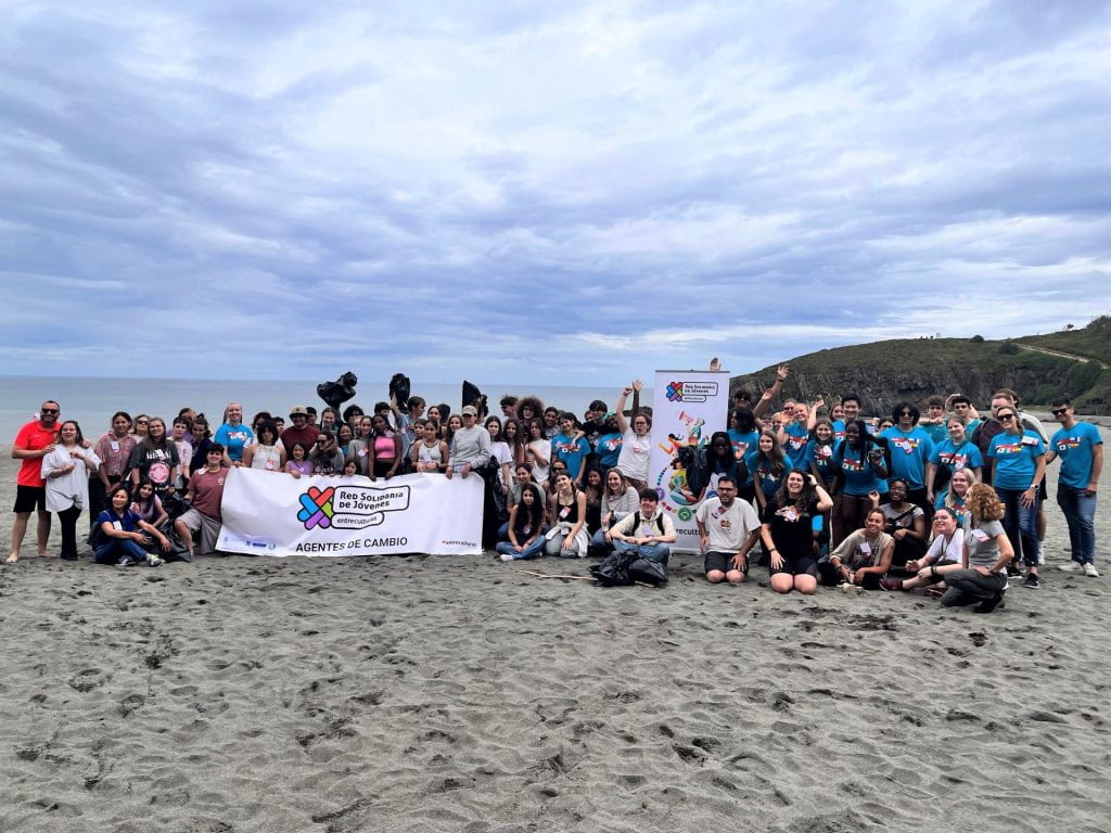 A group of people stand together on beach on an overcast day. They are holding full trash bags and a banner reading beach clean-up in Spanish.