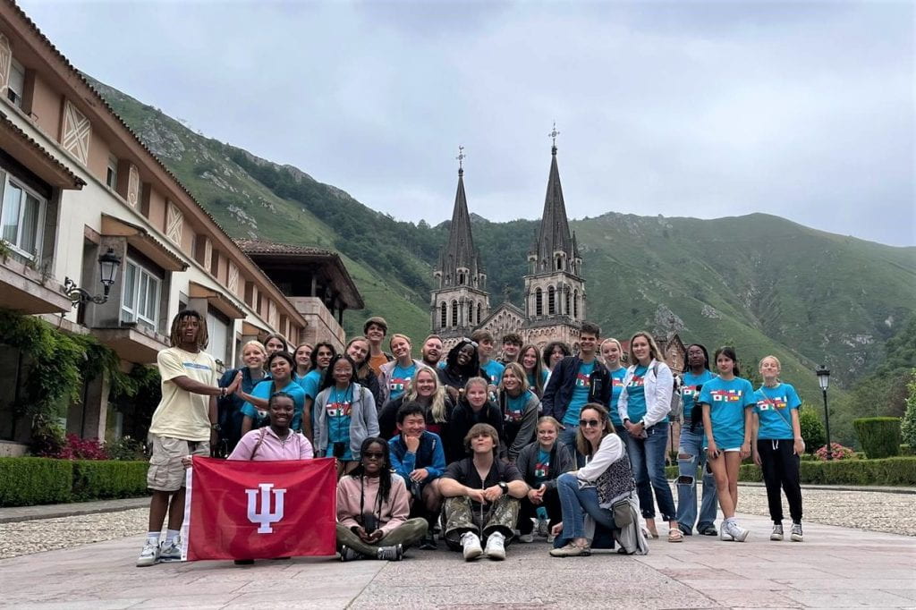 A group of high school students stand in front of a church in Spain holding an IU flag