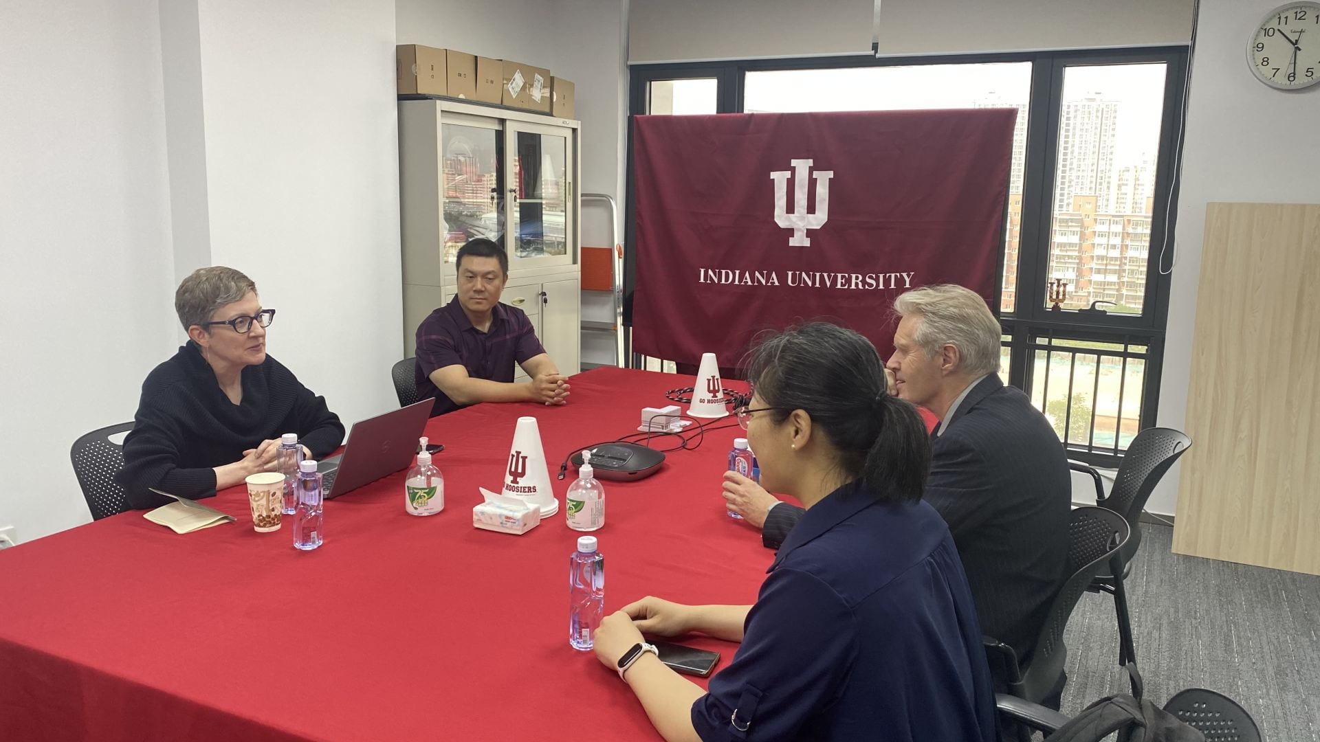 four people sit around a red table. A screen shows the Indiana University trident in the background.