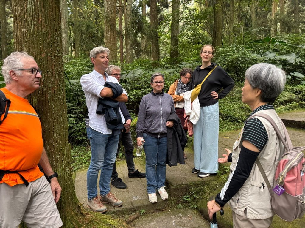 A group of professors stand together in the woods listening to a Taiwanese professor