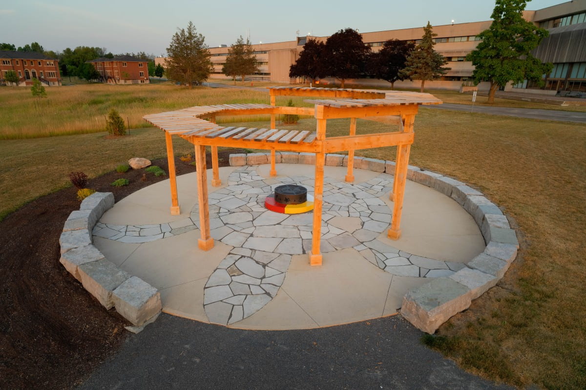 Image is of an outdoor space with a sparse wooden structure and stone seating arranged in a semi-circle around a stone floor containing a turtle pattern and a multi-colored medicine wheel design.