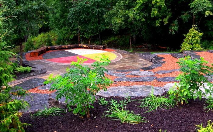 Image is of an outdoor space. Stones for seating are arranged in a tiered semi-circle, facing a large, multi-colored stone in the design of a medicine wheel.