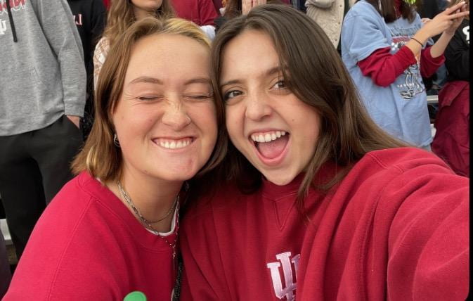 Two students wearing crimson IU sweatshirts make goofy smiles for the camera. One's eyes are closed, and the other has her armed wrapped around her friend's shoulder.