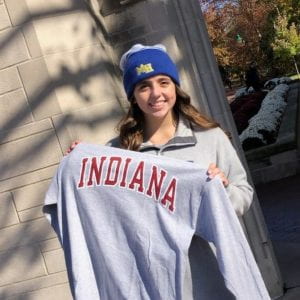 Student in blue stocking camp poses with Indiana sweatshirt in front of IU's limestone Sample Gates.