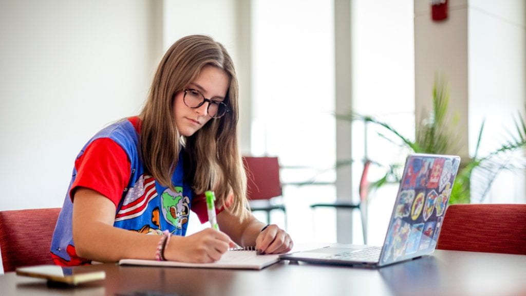 Studious female student in red and blue t-shirt sits at table with laptop and notebook.