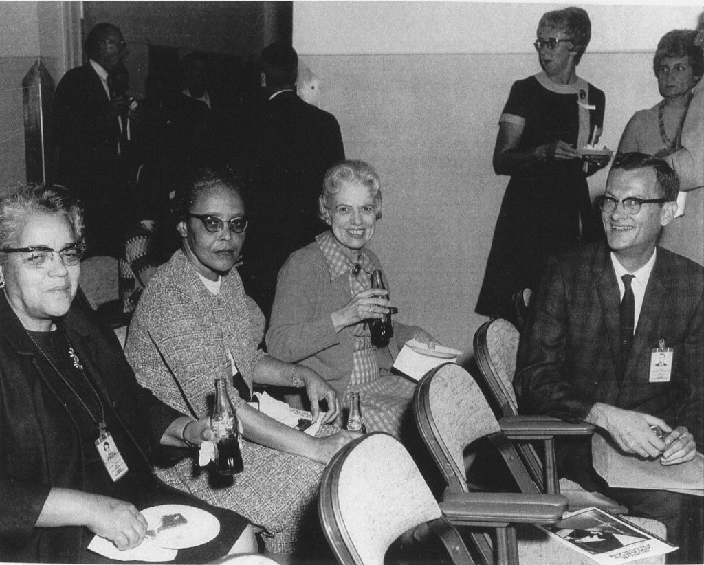 An original black and white photograph of Dorothy Vaughn sitting with two other women and one man. All are dressed in business clothing and wear badges, likely those required while working at NASA. Each is drinking a glass bottle of soda and smiling. Two other women and three men stand in the background, talking to one another.