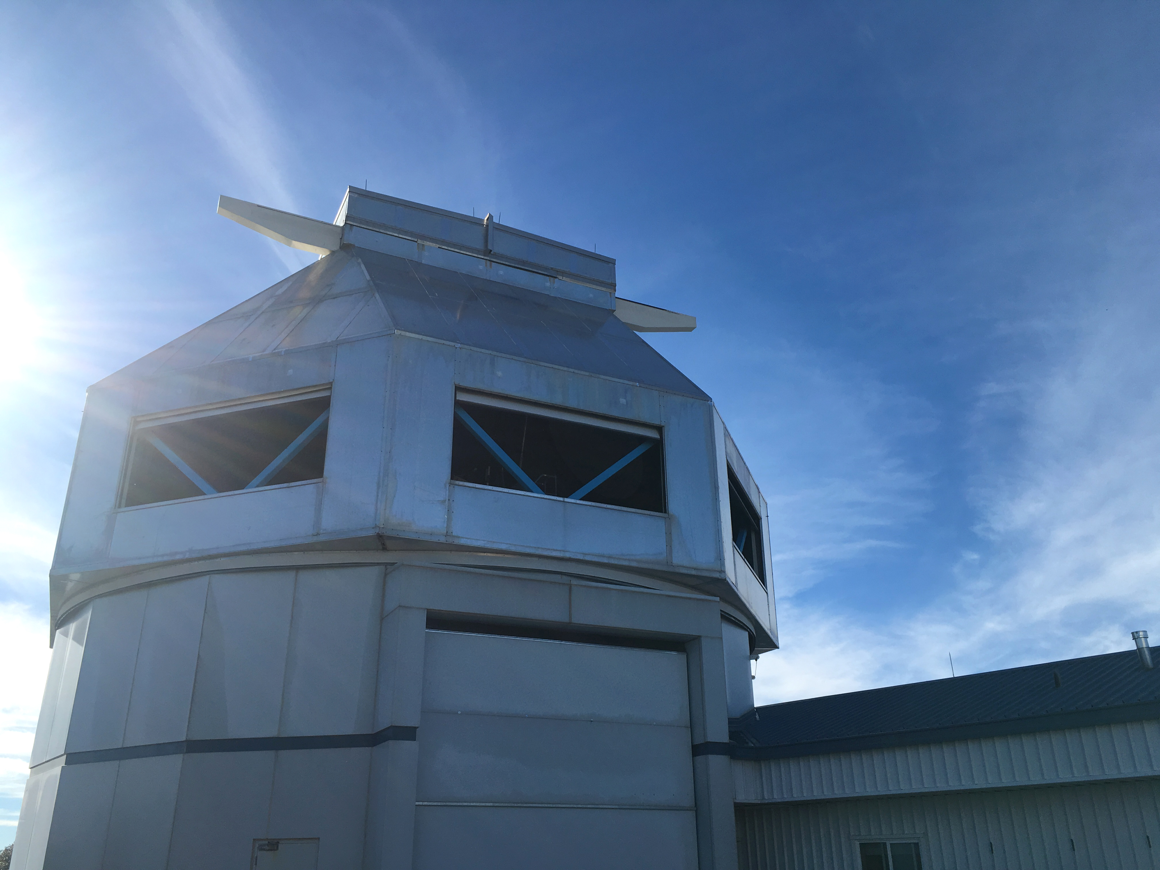 Looking up at a large silver building, vaguely octagonal in shape, that contains the WIYN telescope. There is a blue sky behind it.