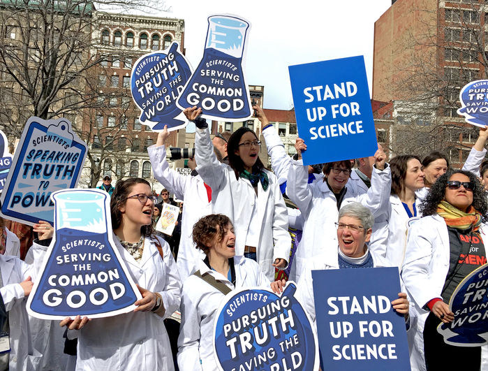 Science activists wearing white lab coats hold signs such as “Scientists serving the common good,” “Stand up for science,” and “Scientists speaking truth to power.” They are gathered at a rally in Boston to “Stand Up for Science.” Many of the rally participants were also attendees at the AAAS annual conference during the same afternoon.