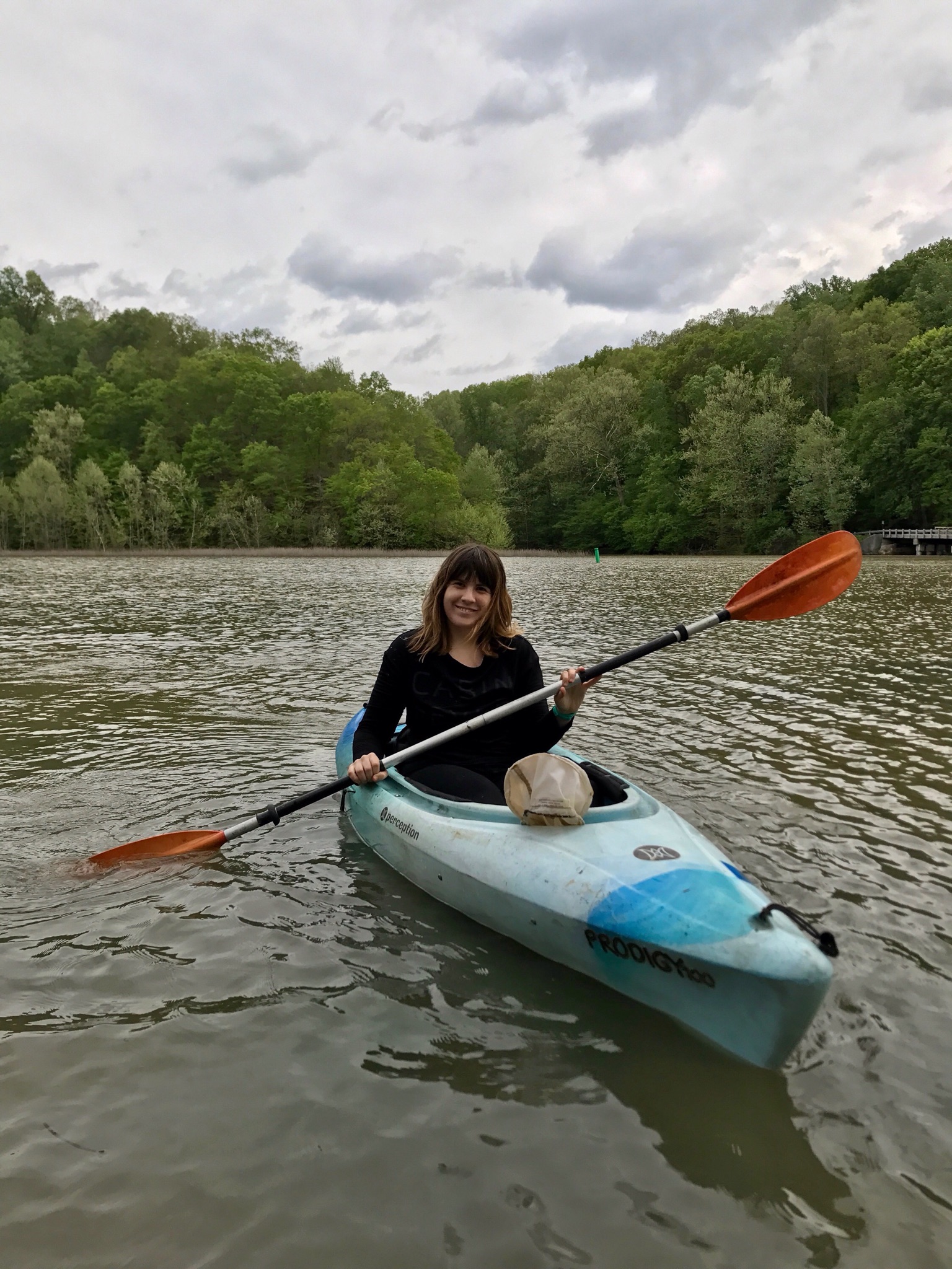 A women sits in a blue boat (kayak) holding a double-sided paddle in the middle of a lake. Trees and cloudy background.