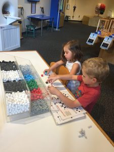 Two children piecing together different colored plastic pieces on a white tabletop to make atom-based chemical structures from a laminated handout on the table. 
