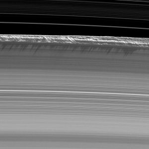 Rings of Saturn are shown in black and white as slightly arced lines across the image. The rings are continuous for most of the image until the structures at the edge of the B ring can be seen jutting up and casting shadows. The top of the image shows ringlets in the division between the A and B rings against the blackness of space.