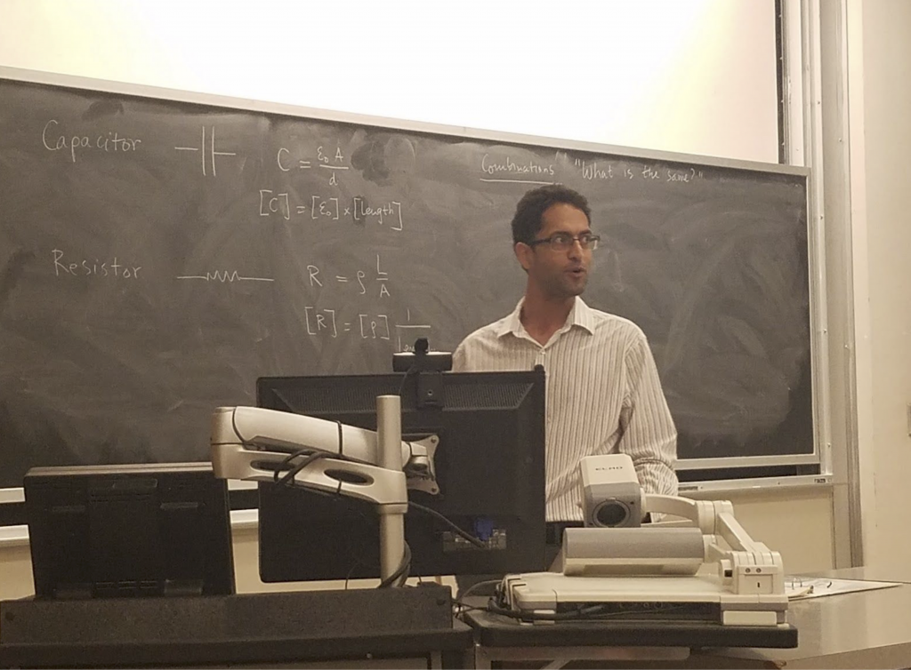 A man stands in front of a chalkboard, where equations are written. There is also a computer on a desk in front of him.