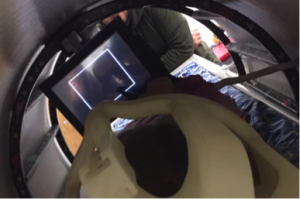 This images shows the view from the child's head within the mock MRI machine toward her feet. In the foreground is the plastic head covering worn to hold her head in place during the scan. In front of her is the tablet, blank at the moment, on which she will write. Past her feet is an adult man leaning on the far edge of the table.