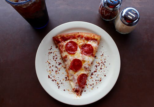 [Slice of pizza on a white plate next to a drink and condiments.]
