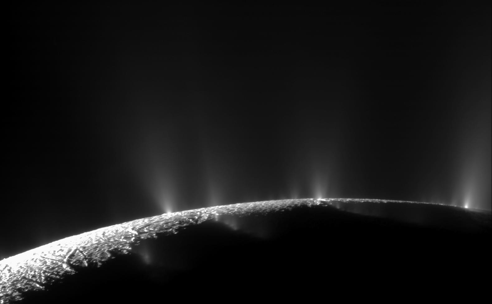 A small section of a darkly lit moon with bright jets of water shooting out from the surface against the blackness of space.