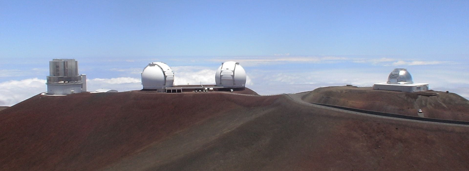 The domes of four telescopes on top of a mountain with the clouds visible below them.