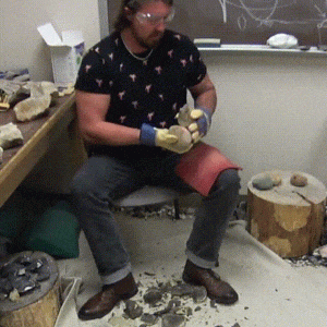 A video of stone toolmaking: a man hits a rock, which he is holding in his left hand in his lap, with another rock, held in his right hand. A sharp tool, called a flake, breaks off of the rock in his lap and falls onto the floor.