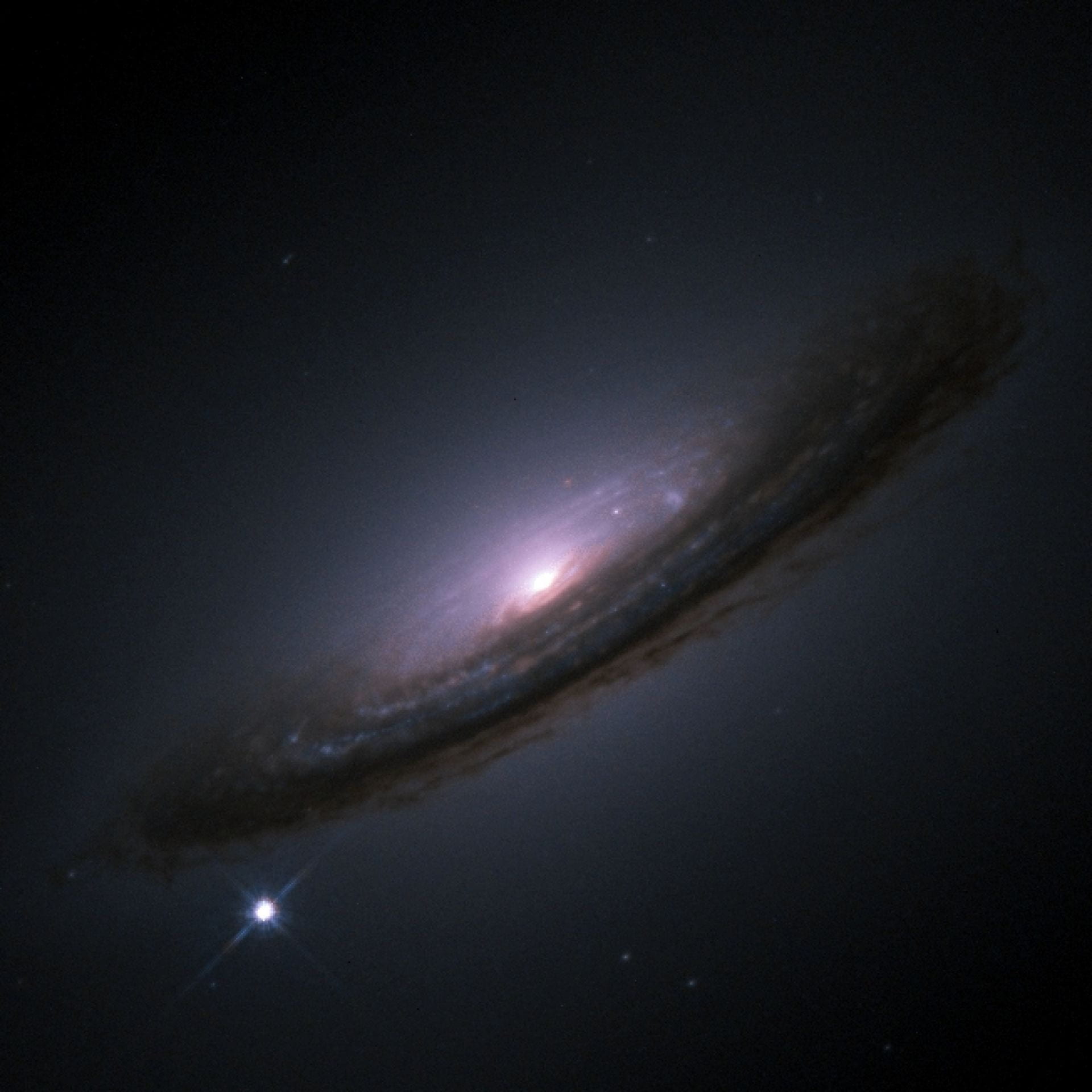 A disk galaxy is seen side-on like a dusty frisbee. Dark clouds are seen against a dark background with a purple glow coming out from the center of the disk. In the lower left, a bright white dot shines.