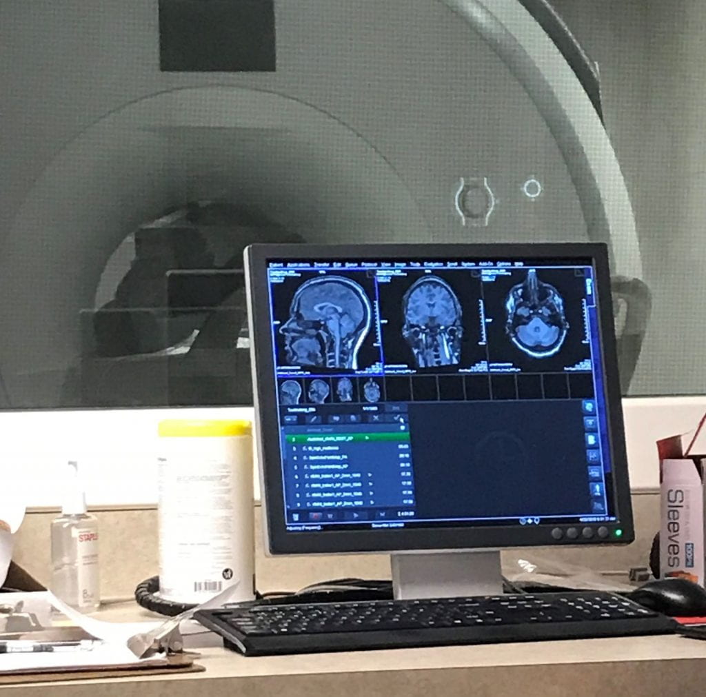 MRI images of a brain in the foreground, a glass windo showing the participant laying in the MRI scanner in the background.