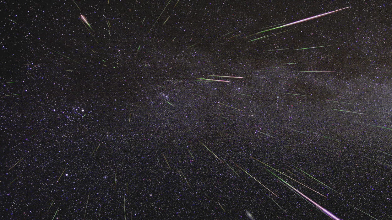 Against a dark sky with many faint background stars, streaks of light radiate outward from the upper middle of the image. Most of the streaks - meteors - are white, but a few have hints of green or purple in their light trail. 