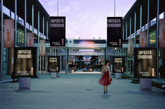 A woman in a red dress and black high heels is standing in the middle of a cobblestone walkway and is looking down at the ground. She is surrounded by pillars with advertisements for an Armani fragrance, and the phrase “Imposter Syndrome” is written at the top of each advertisement in large capital letters.
