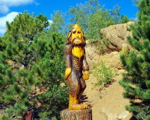 A statue of bigfoot sits in the middle of a forest with trees flanking it on both sides and brown-grayish rocks behind it.