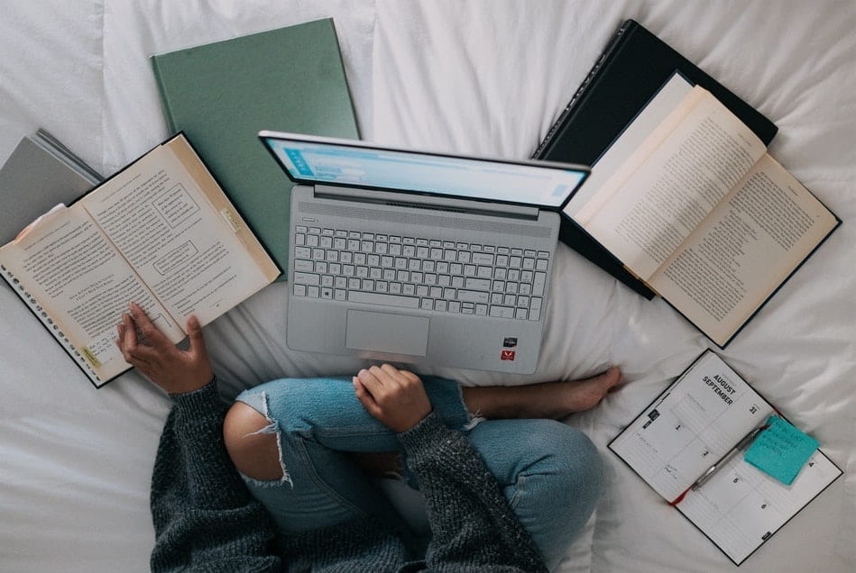 [aerial view of someone wearing jeans who is studying in front of laptop sitting criss-crossed with several books laid to their side against white sheets]