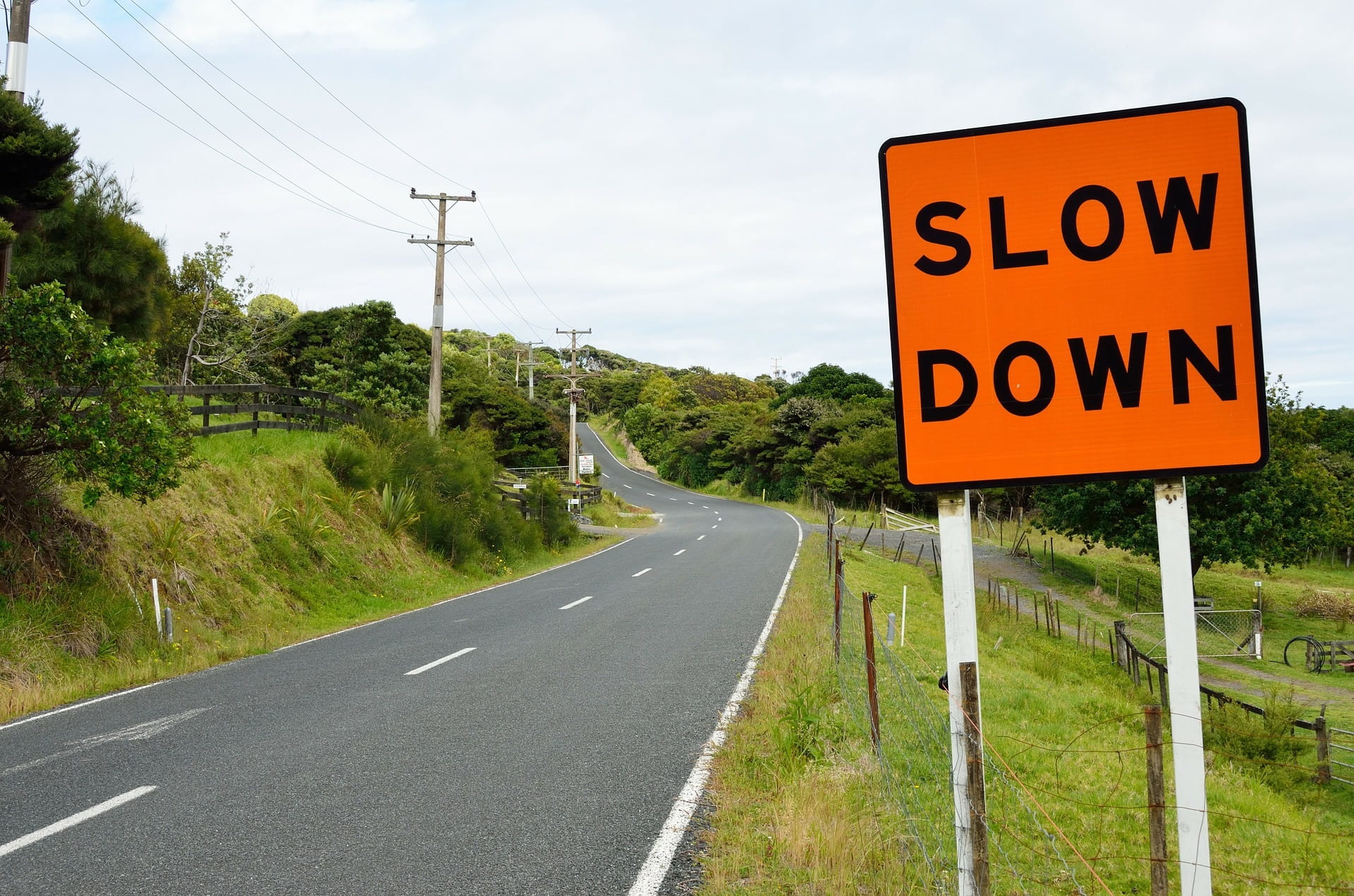 Image of a winding rural road with an orange and black road sign with the words “slow down”.