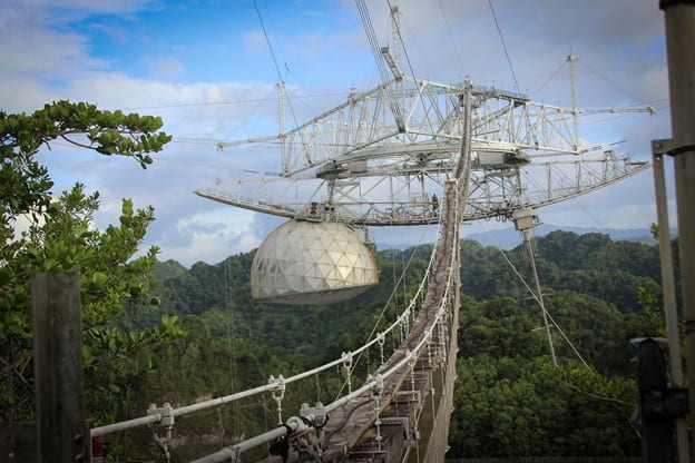 The receiver for the Arecibo telescope. A large white half dome is suspended in the air by a complex set of wires and struts. Behind the receiver the Puetro Rican forests can be seen.