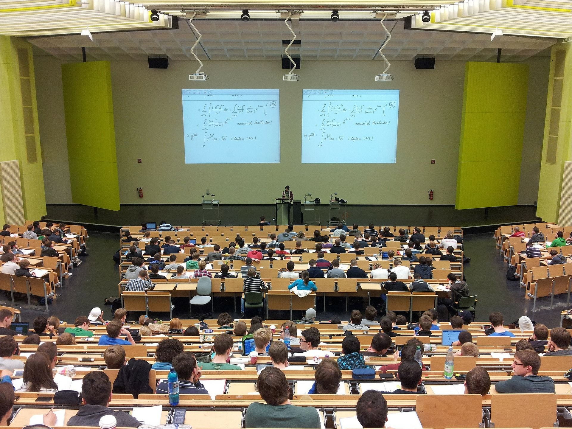 Pictured is a crowded university lecture hall, with students looking at math equations on the overhead projection and a single lecturer at the front of the room