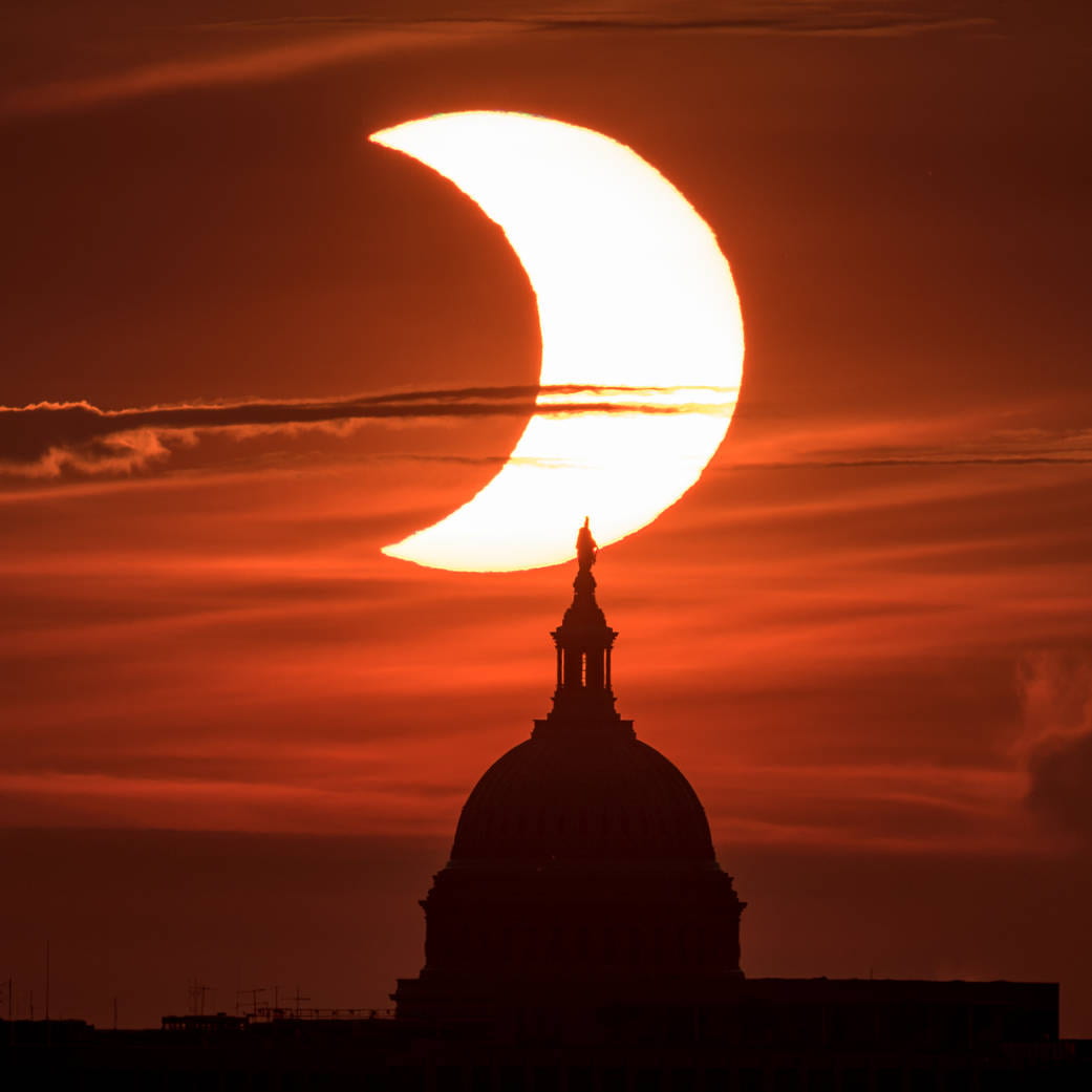 [The sun rises behind the United States Capitol Building during a partial solar eclipse.]
