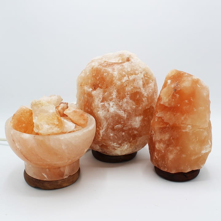 Three pinkish salt lamps with wooden bases against a white background. The leftmost one has been polished into a bowl that is full of pinkish salt crystals.
