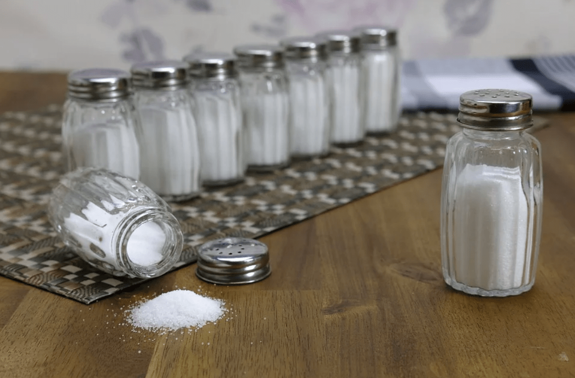 [several bottle of white table salt sit on a metal rack. one of the bottles has spilled over on to the table. .