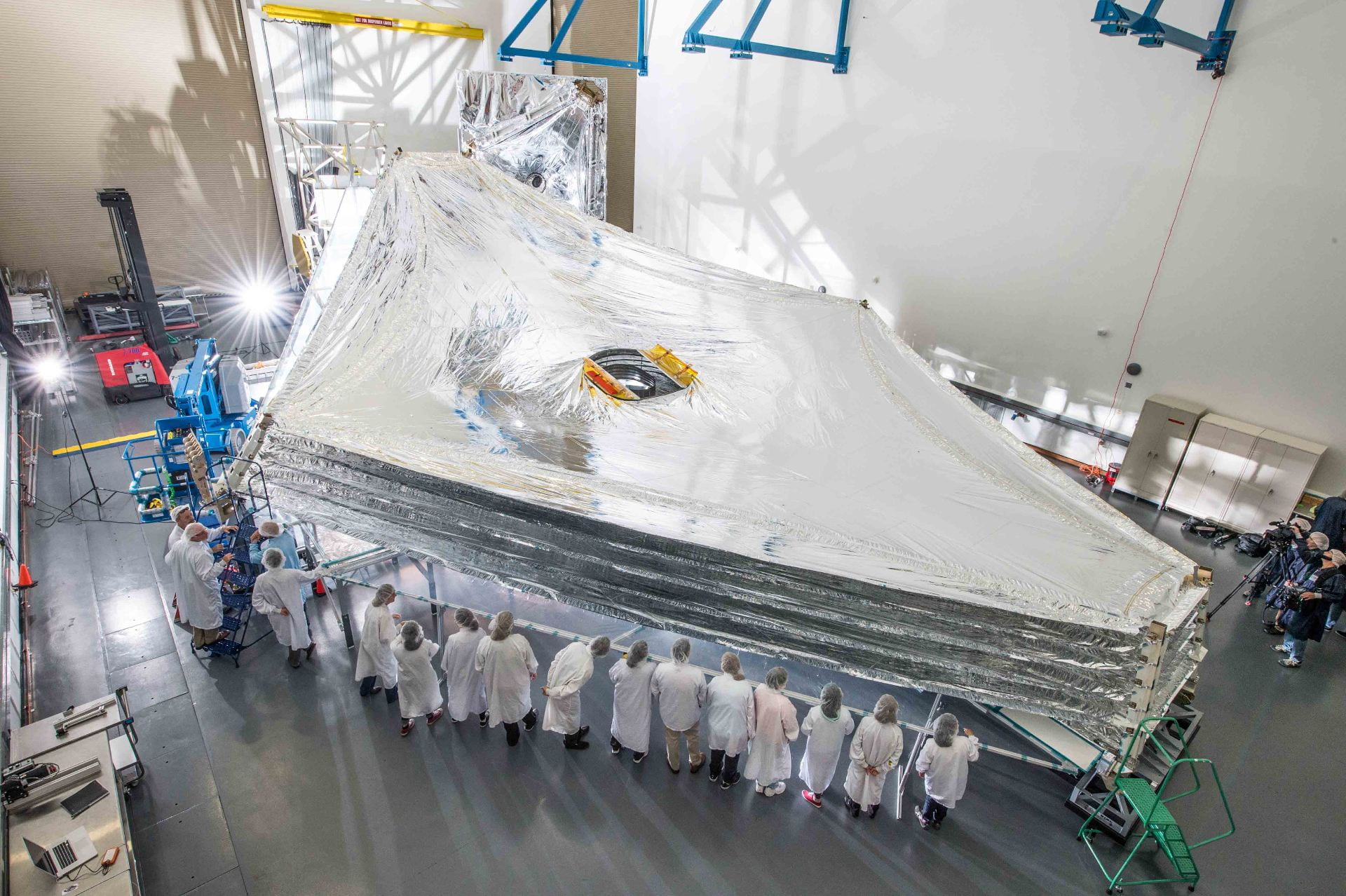 A large silver sunshield is unfolded while a bunch of scientists look on. The shield is approximately diamond-shaped and one side has twelve people standing alongside it.