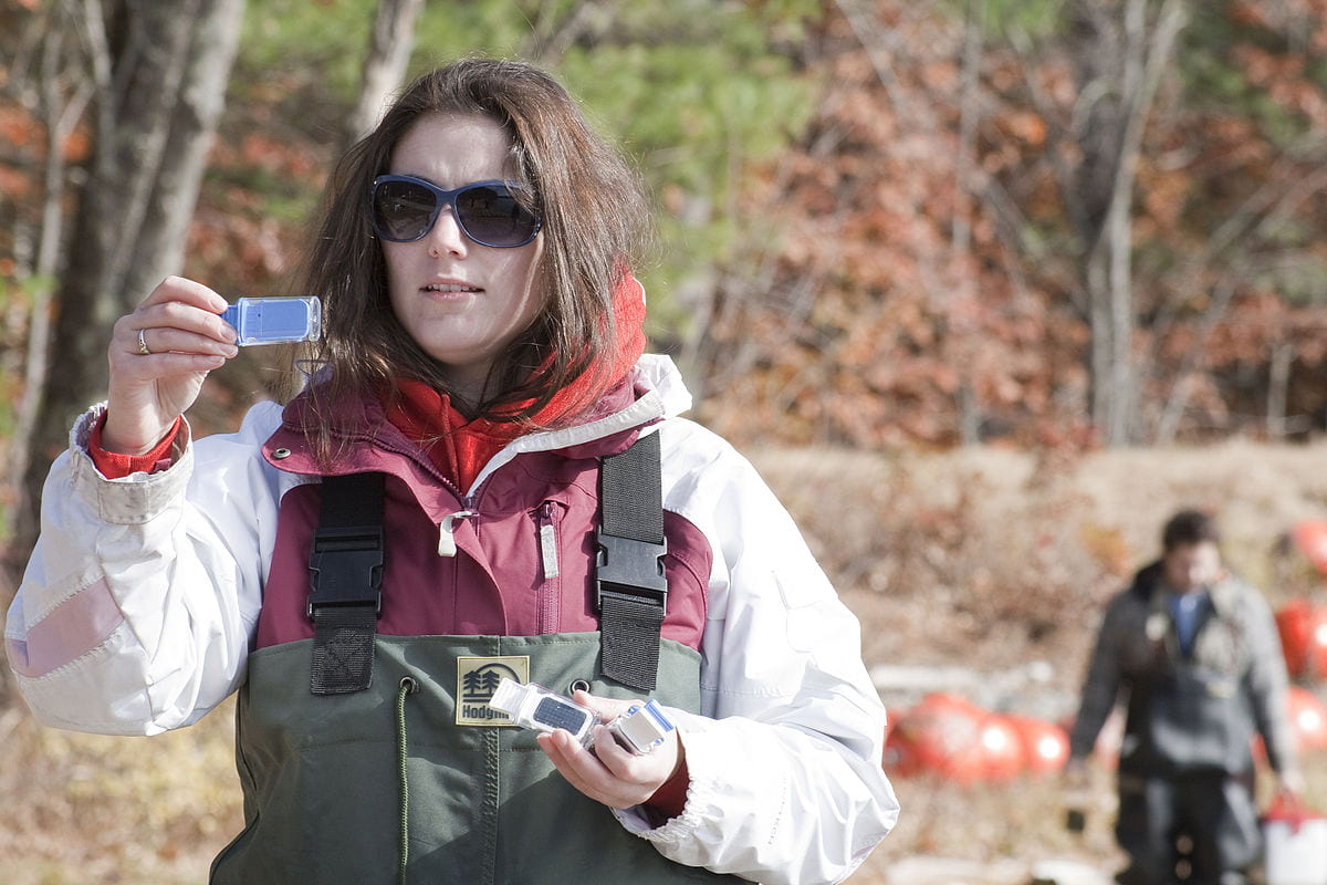 A female scientist in waders views a blue science test outdoors on a fall day.