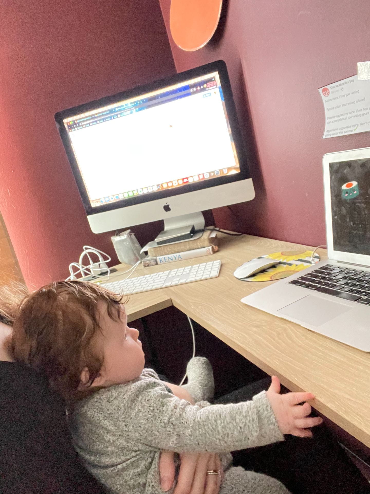 A four month old infant stares at a laptop playing a video of dancing cartoon fruit while schoolwork is visible on an iMac on the other half of the desk