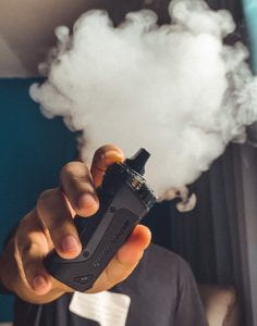 A person is holding a large vape in the front of the photo, and in the background there is a large puff of smoke.