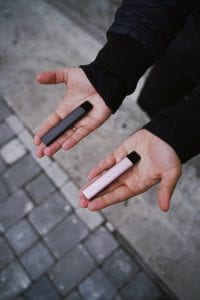 A hand holding 2 vapes, one in each hand, and the vapes resemble a USB flash drive. One is pink and the other is black.