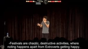 Image shows a man on a stage with a mic. Image also shows closed captions that read "Festivals are chaotic, destructive activities where nothing happens apart from extroverts getting happy"
