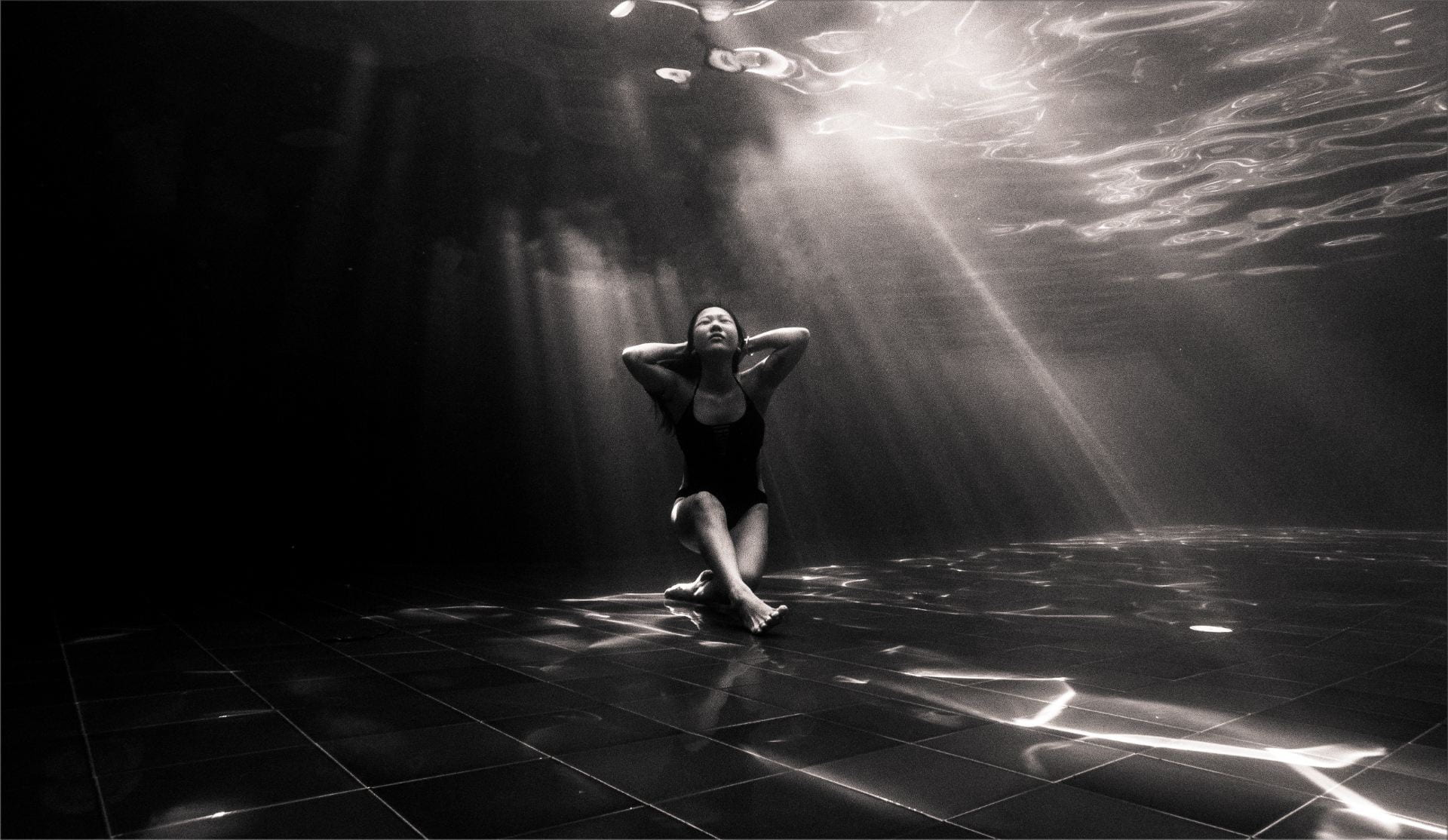 [Black and white photo of a person sitting underwater at the bottom of a pool with light streaming in behind them.]