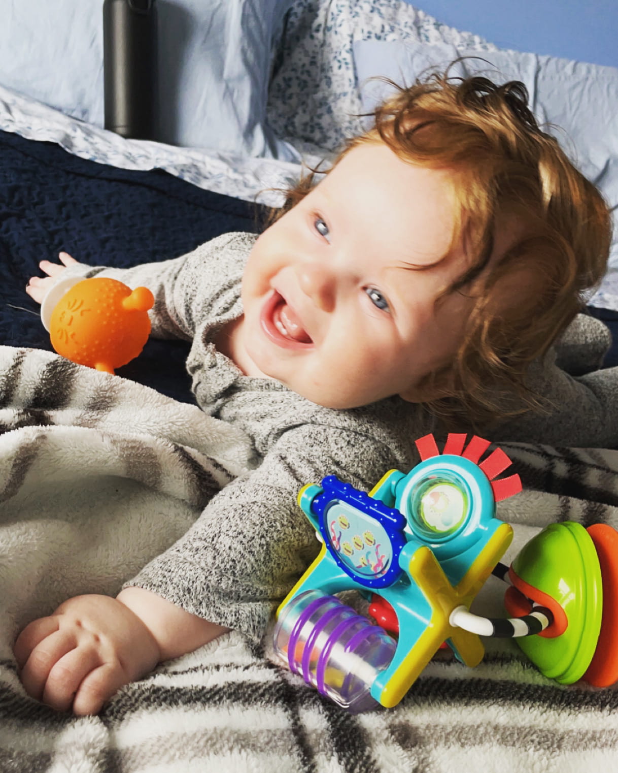 [Pictured is a 4-month-old baby laying on his stomach on a bed while smiling at the camera and playing with a rattle toy and a teething toy.]