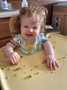 A six month old baby sitting in a high chair with a blue bib on. He has food remnants on his cheeks and bits of food spread out on the high chair tray.