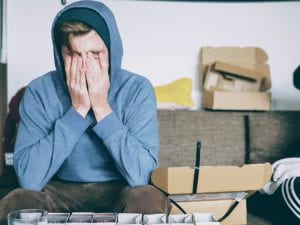 young adult wearing hooding sitting in a room with open boxes. Person has elbows on knees and is covering face with hands.