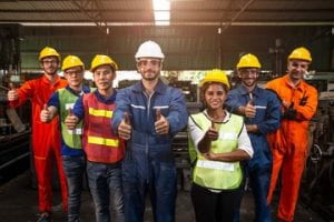 Six male and one female workers of different races in a warehouse wearing hardhats and giving a thumbs up.
