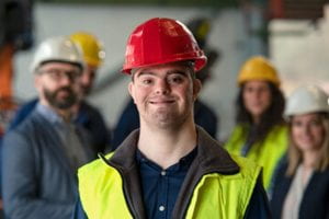 Young worker with Down Syndrome wearing a yellow vest and red hard hat as he stands with coworkers.
